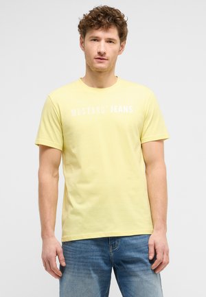 Junger Mann mit lockigem Haar, der ein gelbes "Mustang Jeans" T-Shirt und hellblaue Jeans trägt, steht vor einem schlichten Hintergrund.