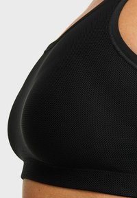 Närbild av en person som bär en svart texturerad sportbh, vilket visar tygdetaljer och passform på övre delen av kroppen mot en enkel bakgrund.