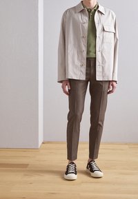 Béžová bunda s límečkem a přední kapsou, vrstvená přes zelené polo tričko. Hnědé slim-fit kalhoty a černé tenisky s bílými akcenty dokončují outfit.