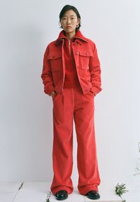 Veste en velours côtelé rouge avec poches avant portée sur un top en tricot rouge assorti et un pantalon large, accompagnée de chaussures noires.