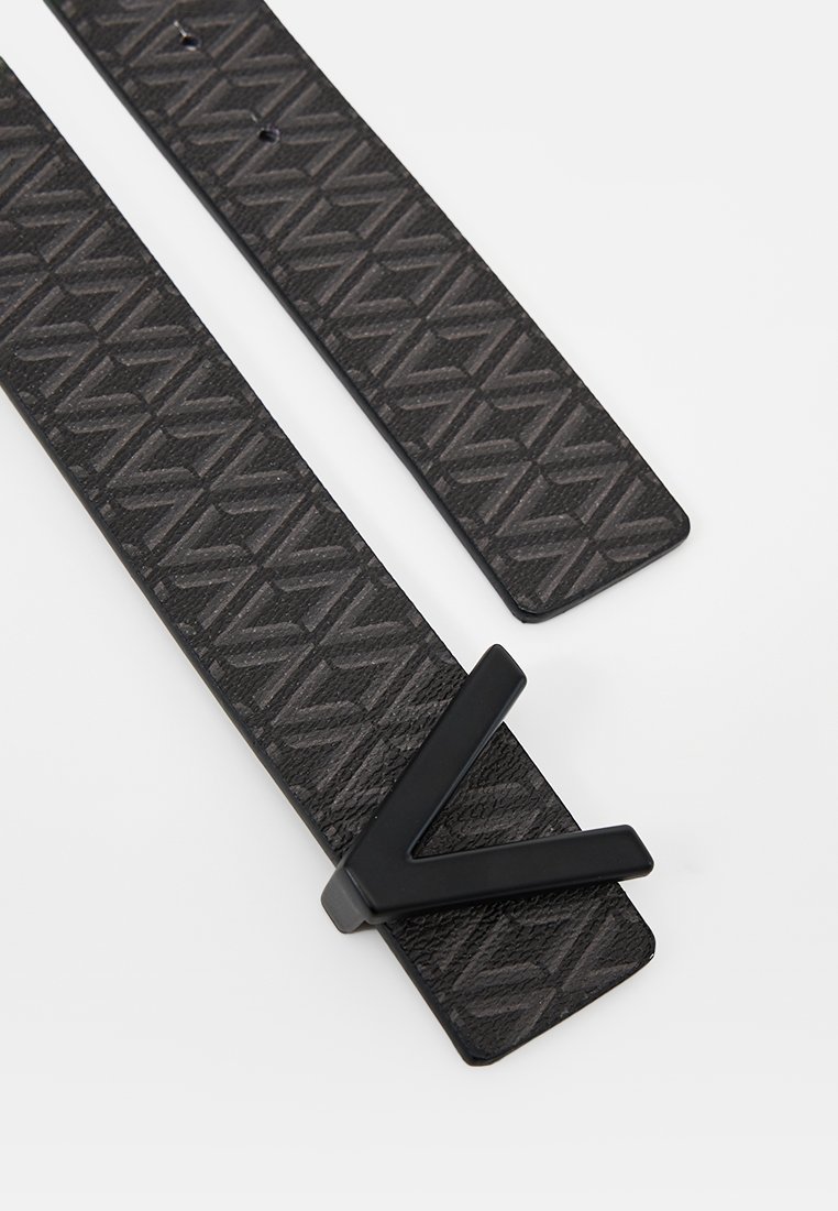 Ceinture noire à motifs avec texture géométrique, dotée d'une boucle en V noire. Deux sections de sangle visibles, dont une avec des trous pour le réglage.