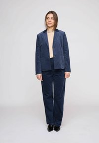 Completo giacca e pantaloni in velluto a coste blu navy, con collo a risvolto, due tasche frontali e vestibilità leggermente ampia. Tessuto strutturato con righe verticali.