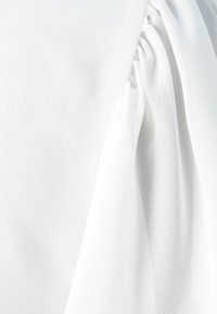White blouse fabric featuring soft, textured material with gathered detail on the shoulder, creating gentle draping.