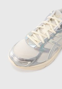 Lekki but sportowy z siateczkową cholewką w kolorze kremowym z metalicznymi srebrnymi akcentami, wyposażony w płaskie sznurowadła i amortyzowaną podeszwę.