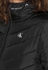 Doudoune noire avec un design matelassé horizontal, fermeture éclair et un logo "ck" blanc sur la poitrine gauche. Tissu en nylon lisse.
