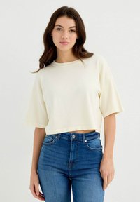 Camiseta corta de color crema con cuello redondo y mangas cortas, combinada con jeans de mezclilla azules ajustados, mostrando una textura suave.