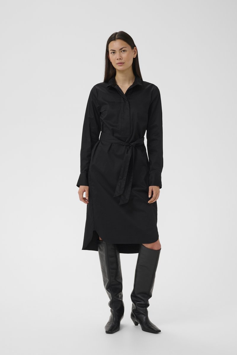 Hosszú sötét hajú nő, fekete, öves, hosszú ujjú ingruhában és térdig érő fekete csizmában, egyszínű világos háttér előtt állva.