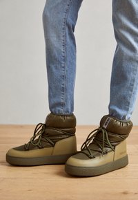 Bottines rembourrées vert olive avec semelle en caoutchouc, dotées de lacets et d'une base texturée. Conçues pour la chaleur et le confort, avec un profil épuré.