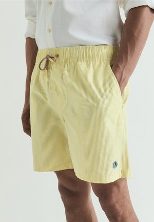 Homme portant un short de bain rayé jaune et blanc avec une taille à cordon et un petit logo rond près de l'ourlet sur un fond clair.