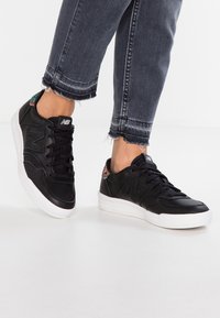 Baskets en cuir noir avec une semelle en caoutchouc blanche, présentant un design texturé et un accent floral sur le talon. Lacées avec des lacets noirs.