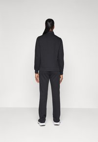 Sweatshirt noir et pantalon de jogging assorti en tissu doux, avec une coupe décontractée et des poignets côtelés, portés avec des chaussures de sport blanches.