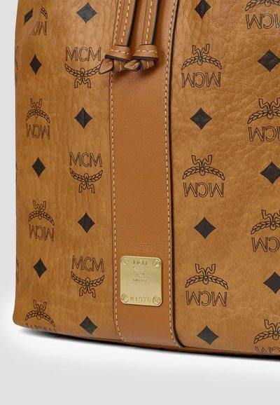 Sectie van een MCM-tas van tan leer met zwart logopatroon, een centrale leren strook met gebonden riemen, en een gouden metalen merkplaatje met serienummer.