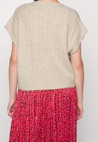 Pull en tricot beige avec manches courtes retroussées et motif en câble. Associé à une jupe plissée rouge avec de petites imprimés floraux et des pois contrastants.