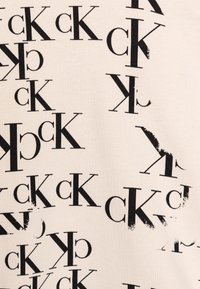 Krémová textilie s opakovaným černým potiskem písmen "CK" v různých orientacích a velikostech, vytvářející texturovaný, dynamický vzor.