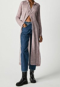 Chemise longue à boutons dans un motif géométrique rouge et blanc, fabriquée en tissu léger. Associée à un jean bleu taille haute et des bottes noires.