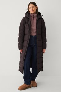 Donker pufferjack met een lange ontwerp, ritssluiting en capuchon. Het heeft gestikte secties en wordt gecombineerd met een lichte coltrui en blauwe jeans.
