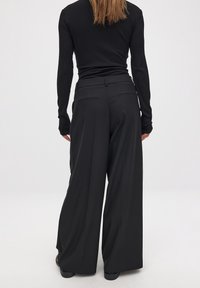 Wide-leg pants in black fabric, featuring a high waist, subtle front pleats, and slanted back pockets, paired with a fitted long-sleeve top.