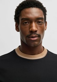 T-shirt nera con colletto a coste beige e cuciture piatte. Primo piano del viso di un modello maschile, che mette in mostra una pelle liscia e capelli corti e ricci.