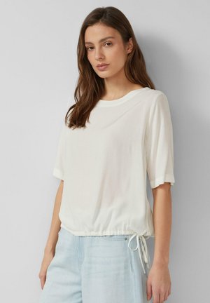 Młoda kobieta z długimi brązowymi włosami, ubrana w luźny biały krótki top z wiązaniem z boku oraz jasnoniebieskie dżinsy, stojąca na szarym tle.