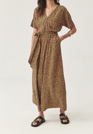 Femme portant une robe portefeuille beige à pois noirs, manches courtes, ceinture nouée à la taille, sandales noires et un bracelet en or au poignet.