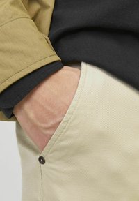Beige strukturierte Hose mit einer Seitentasche und einem dunklen Knopf. Ein olivgrüner Jackenärmel ist teilweise sichtbar, der über einem schwarzen Hemd liegt.