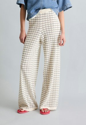 Osoba nosząca luźne kremowe spodnie z granatowymi wzorami w kształcie jodełki, krótki jeansowy top i czerwone klapki, stojąca na jednolitej szarej podłodze.