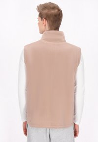 Gilet in pile beige chiaro con colletto alto, caratterizzato da una consistenza morbida e senza chiusure o tasche visibili, indossato sopra una camicia bianca a maniche lunghe.