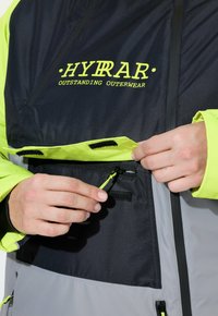 Ręce regulujące neonowożółtą klapę z rzepem nad czarną kieszenią na zamek błyskawiczny na szaro-żółtej kurtce o wysokiej widoczności z napisem "HYRAR OUTSTANDING OUTERWEAR".