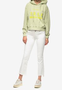Persoon in een lichtgroene cropped hoodie met gele tekst, witte cropped flare jeans en beige-witte sneakers, staande tegen een witte achtergrond.