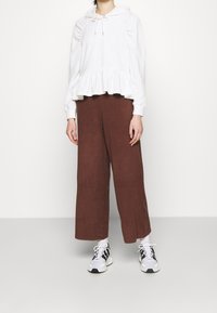 Felpa bianca con orlo ruffle e cappuccio con cordino, abbinata a pantaloni larghi marroni. Il modello indossa sneakers bianche e nere. Texture piatta.