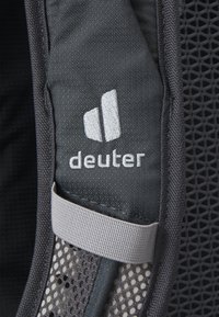 Detaliile rucsacului Deuter includ un material gri, un strat de plasă, o curea gri deschis și un logo brodat cu trei dungi verticale.