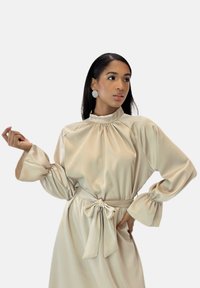 Beige satin kjole med høj halsudskæring, rynkede detaljer og lange, puffede ærmer. Afsluttet med et matchende bånd i taljen og udvidede manchetter.