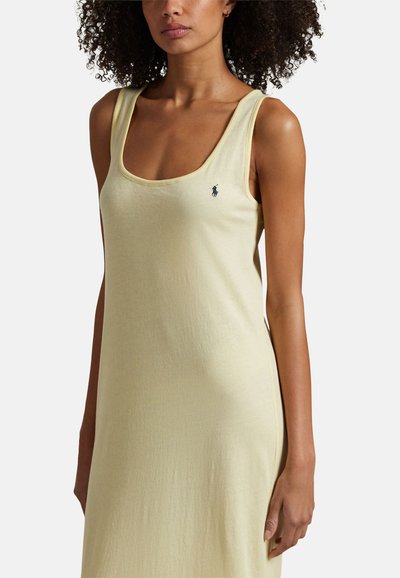 Femme portant une robe sans manches jaune pâle avec un décolleté en U et un petit logo de cheval noir brodé sur la poitrine.