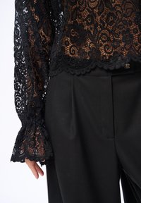 Czarna koronkowa bluzka z długim rękawem w kwiatowe wzory, noszona z czarnymi spodniami o wysokim stanie, ukazującymi częściowo ramię i dłoń.