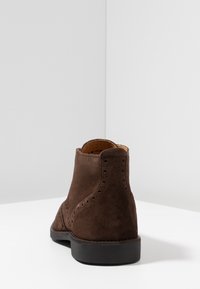 Bottine en daim brun foncé avec perforations décoratives, bout arrondi et semelle en caoutchouc noire, photographiée de dos sur une surface blanche.