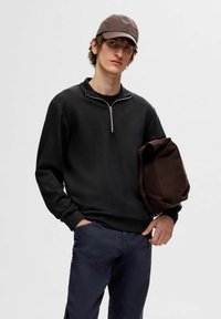 Zwarte sweatshirt met zip en lange mouwen, gecombineerd met donkerblauwe broek. Een bruine tas wordt onder de arm gehouden en er wordt een bruine pet gedragen.