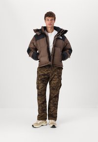 Brun och svart pufferjacka med huva, dragkedja framtill och sidofickor; ihop med kamouflagemönstrade cargobyxor och beige atletiska skor.