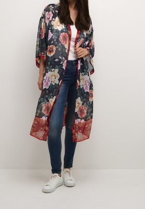 Kobieta ubrana w kwiecisty prześwitujący kimono na białym topie, niebieskich obcisłych dżinsach i białych trampkach, stojąca na jednolitym tle.