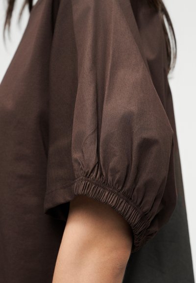 Καφέ πουκάμισο με κοντό μανίκι και ελαστικό σφιγκτήρα στη μανσέτα, που φοριέται στο χέρι, δείχνοντας την υφή του υφάσματος και τη λεπτομέρεια του μανικιού.