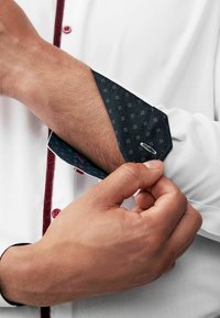 Weißes Hemd mit roter Knopfleiste und dunkel gemustertem Manschette. Die Manschette zeigt ein kleines, sich wiederholendes geometrisches Design und sichtbare Nähte.
