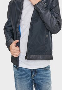 Henkilö, jolla on päällään musta nahkatakki valkoisen paidan ja sinisten farkkujen päällä, pitää takin vetoketjusta yhdellä kädellä suljettuna ja toinen käsi on lähellä kaulusta.