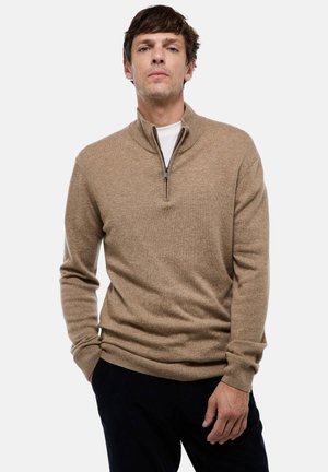 Homme portant un pull zippé beige par-dessus une chemise blanche, les mains dans les poches de son pantalon sombre, se tenant devant un fond clair et uni.