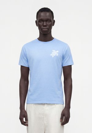 Mand iført en lyseblå kortærmet T-shirt med en hvid skildpaddegrafik på venstre bryst og hvide bukser, stående mod en ensfarvet baggrund.