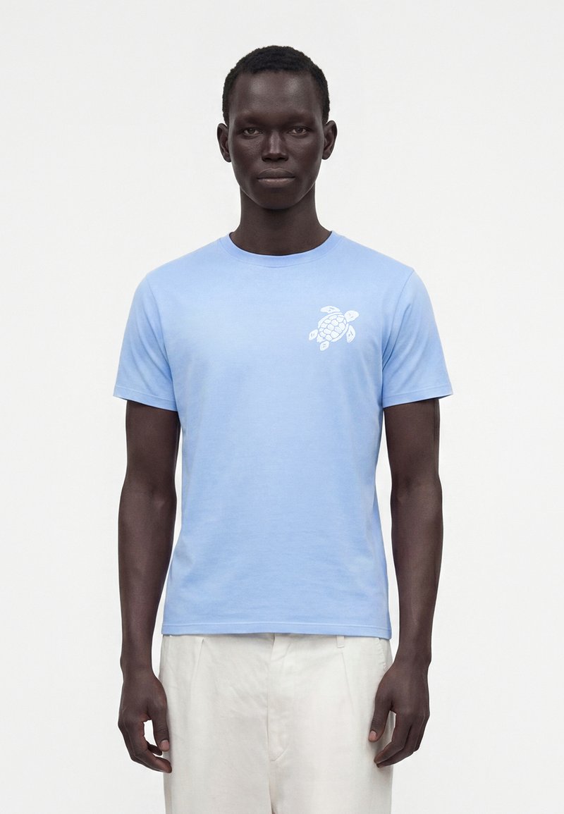 Homme portant un t-shirt bleu clair à manches courtes avec un motif de tortue blanche sur le côté gauche de la poitrine et un pantalon blanc, debout devant un fond uni.