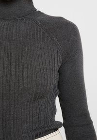 Pull à col roulé en maille côtelée gris foncé avec manches longues, porté rentrée dans un pantalon écru.