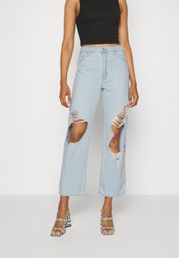 Jeans largos de cor azul clara, com grandes desgastes nos joelhos. Apresenta um fecho de botão e cinco bolsos. Usados com saltos preto e branco com padrão.