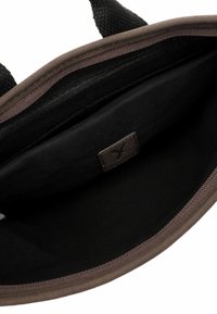 Interior de una bolsa de tela marrón con un forro negro liso, un bolsillo con cremallera y una pequeña etiqueta marrón en la base. Asas texturizadas unidas.