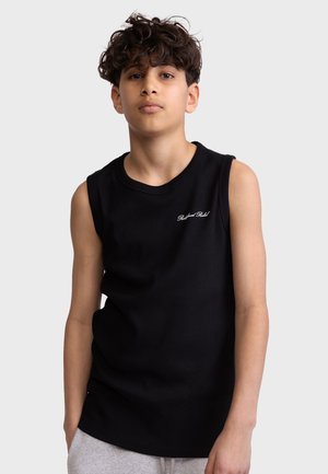 Tienerjongen met krullend haar die een zwart mouwloos shirt draagt met kleine witte tekst op de borst en lichtgrijze broek.
