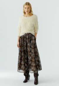 Frau trägt einen cremigen, flauschigen Pullover, einen langen schwarzen Rock mit silbernen Sternmuster und dunkelbraune, hochhackige Stiefel. Sie steht vor einem schlichten Hintergrund.