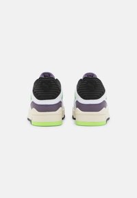 Zapatillas deportivas con parte superior blanca, malla negra y detalles en morado, con un cuello acolchado, suela intermedia verde y suela texturizada para mejor agarre.
